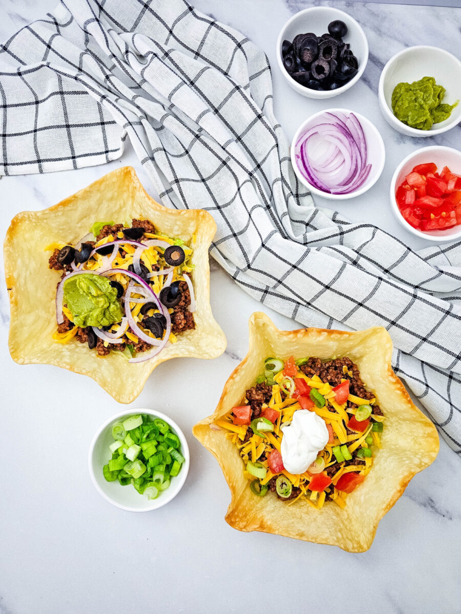 Two ground beef taco salad bowls topped with sour cream and guacamole, surrounded by small bowls of chopped vegetables and black olives.