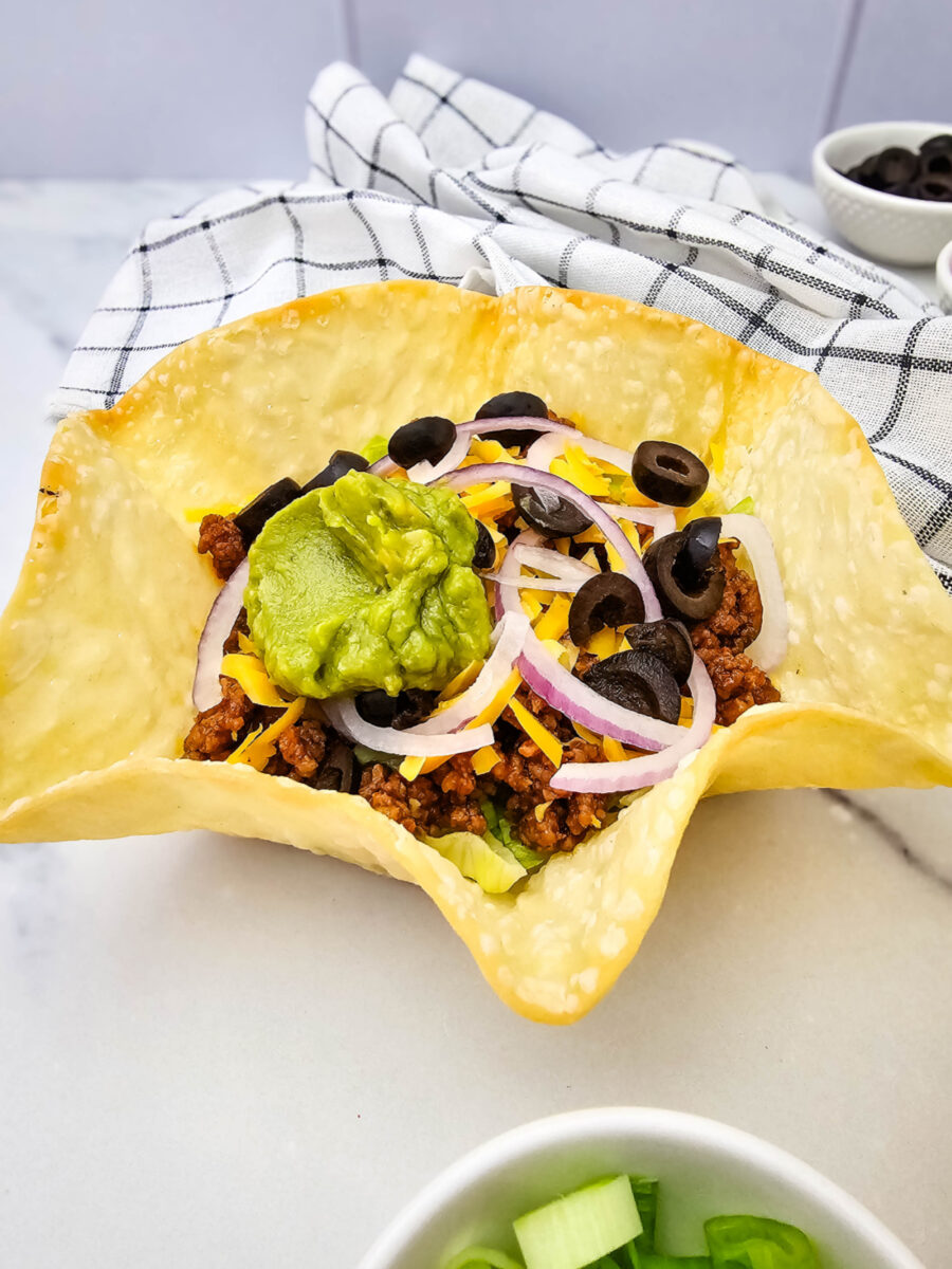 Ground beef taco salad in a baked tortilla shell with lettuce, cheddar, sliced black olives, red onion and a dollop of guacamole.