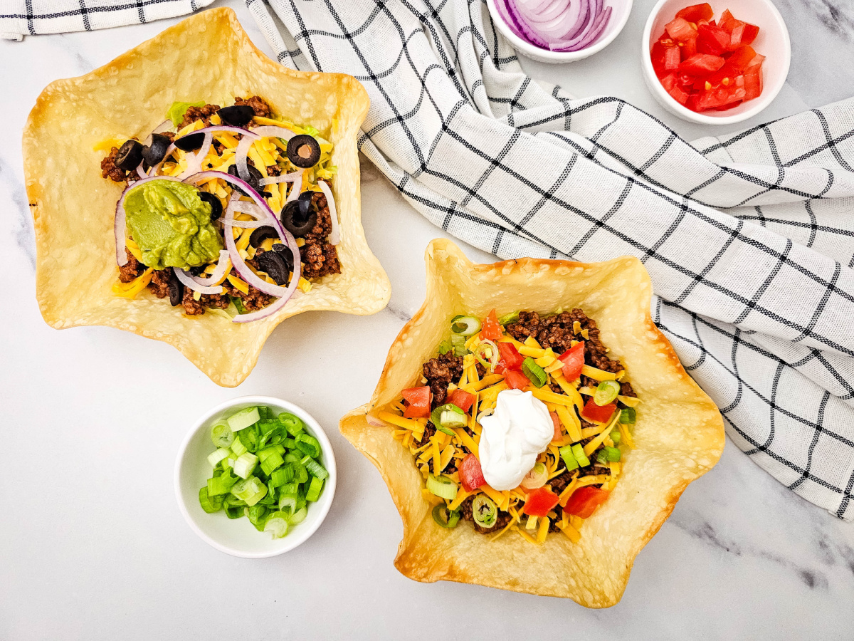 Two taco salads in separate baked tortilla shells, one with guacamole, olives and red onion, the other with sour cream, tomatoes and green onions.