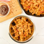 Overhead view of BBQ chicken pasta in a stainless steel bowl next to a cast iron skillet and a cutting board with barbecue sauce and red pepper flakes.