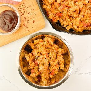 Overhead view of BBQ chicken pasta in a stainless steel bowl next to a cast iron skillet and a cutting board with barbecue sauce and red pepper flakes.