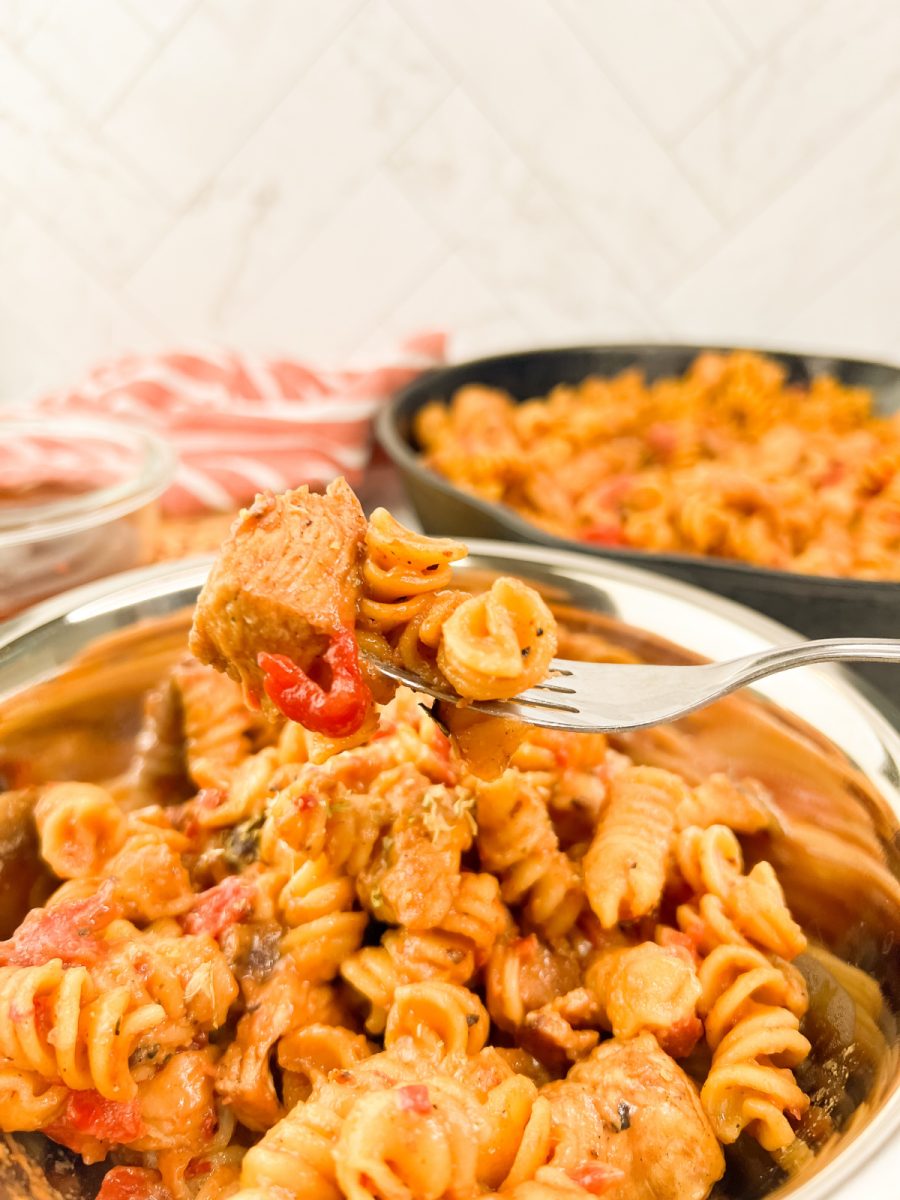 Close-up of a fork lifting a bite of BBQ chicken pasta with visible rotini, chicken, and tomato; bowl and skillet of pasta in the background.