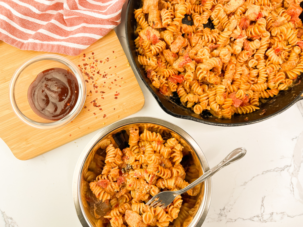 A top-down view of One Pan BBQ Chicken Pasta in a black cast iron skillet and a silver bowl with a fork. A small bowl of BBQ sauce with red pepper flakes is on a wooden cutting board next to a red and white striped towel.