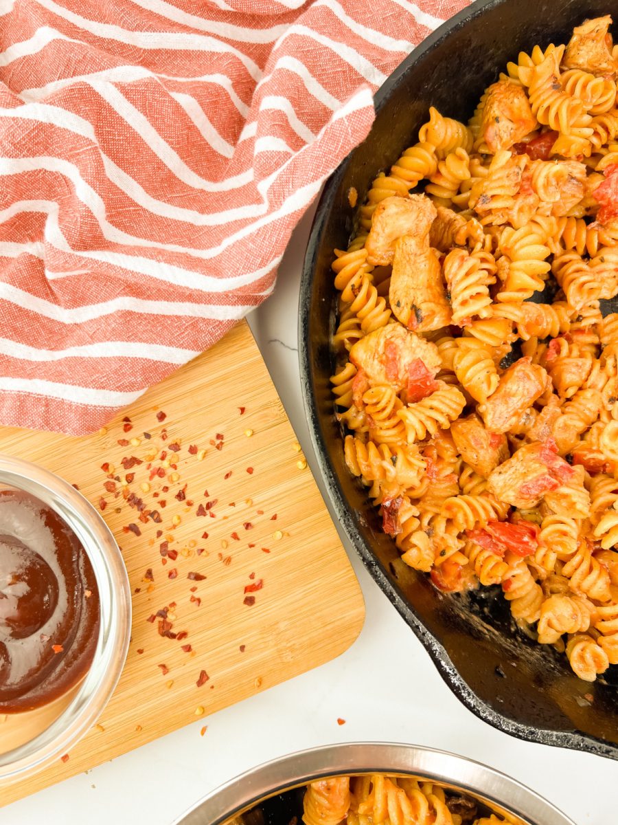 A close-up, overhead view of One Pan BBQ Chicken Pasta in a cast iron skillet. Next to it, a wooden cutting board holds a small bowl of BBQ sauce and scattered red pepper flakes, with a red and white striped towel.
