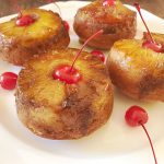 A square white platter holds four jumbo pineapple upside-down muffins, each crowned with a maraschino cherry with the stem, surrounded by extra cherries.