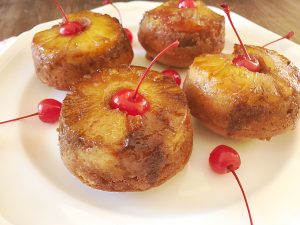 A square white platter holds four jumbo pineapple upside-down muffins, each crowned with a maraschino cherry with the stem, surrounded by extra cherries.