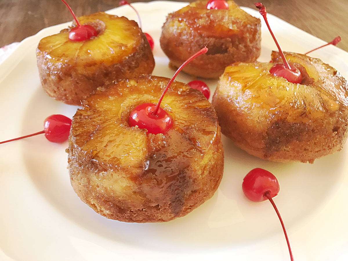 A square white platter holds four jumbo pineapple upside-down muffins, each crowned with a maraschino cherry with the stem, surrounded by extra cherries.