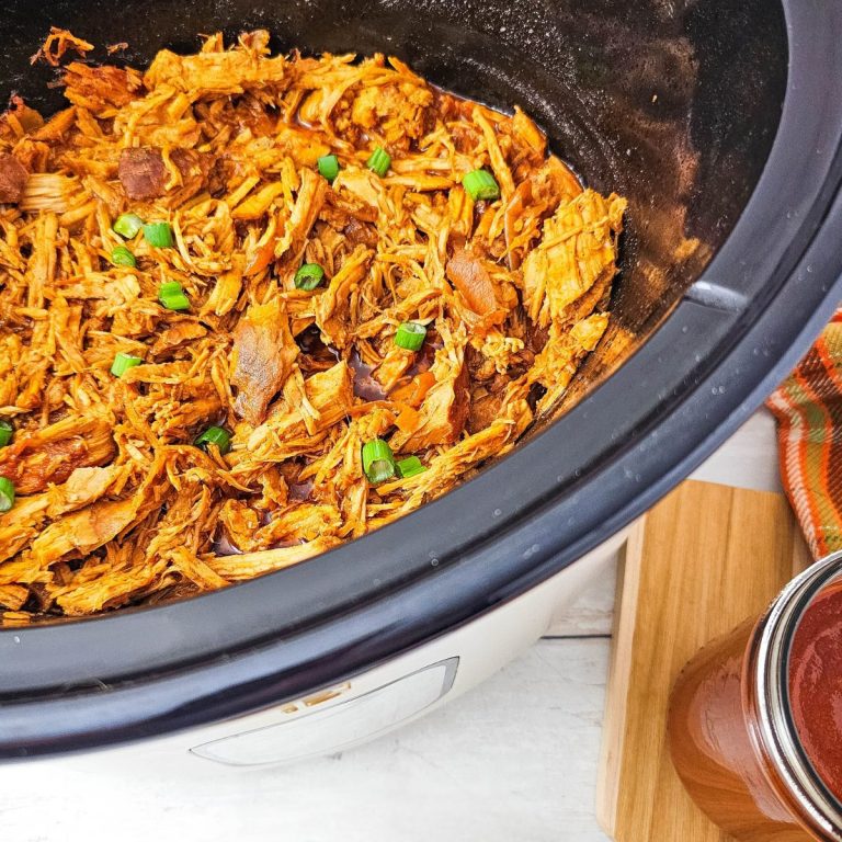 A close-up shot shows a black crock pot filled with shredded BBQ pulled pork, garnished with chopped green onions. The pulled pork appears tender and coated in a rich, reddish-brown sauce. To the right of the crock pot, a clear glass jar containing more of the same BBQ sauce is visible, sitting on a light-colored surface next to a partially seen orange and white plaid cloth.
