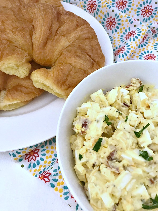 Bacon Cheddar Egg Salad in white bowl.. A plate of fresh crossiants are stacked beside the egg salad