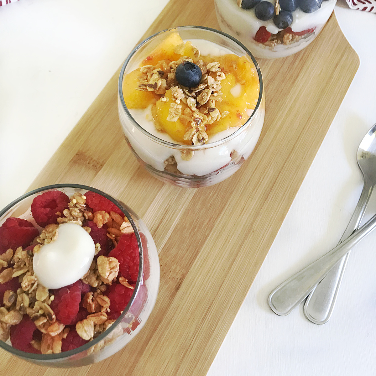 Three glass bowls filled with yogurt parfaits on a wooden cutting board. Each parfait is layered with yogurt, fresh fruit (raspberries, blueberries, peaches), and granola.