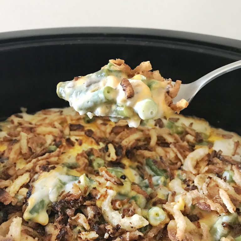 Close-up image of a slow cooker filled with cheesy green bean casserole topped with crispy fried onions. A spoon lifts a portion, showing tender green beans coated in a creamy, melted cheese.