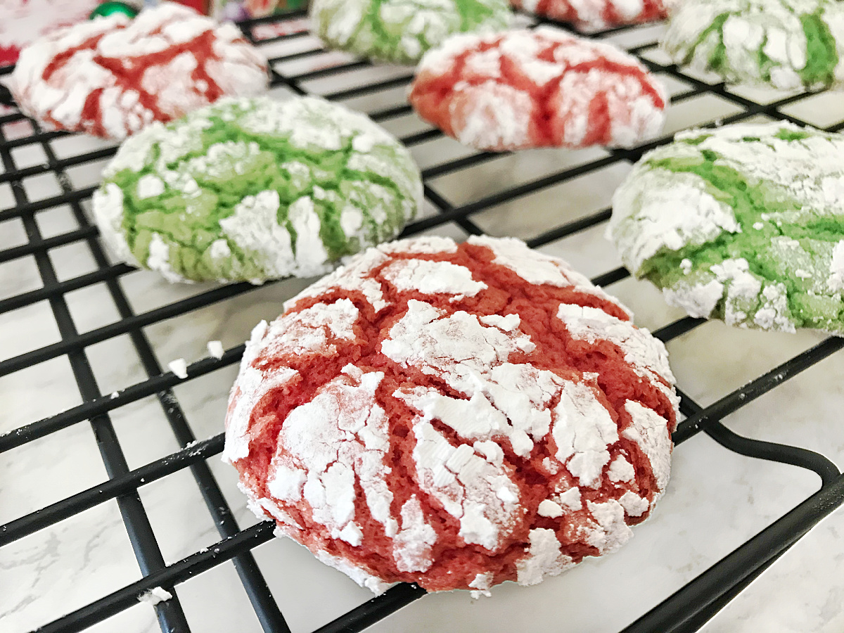 Extreme close-up of a red Christmas Crinkle Cookie showing the deep cracks and heavy powdered sugar coating, with blurred green and red cookies on a cooling rack in the background.