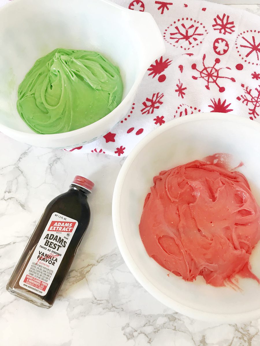 Two white bowls containing bright green and red cookie dough with a bottle of vanilla extract next to them, ready for baking.