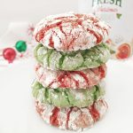 Stack of four baked red and green Christmas Crinkle Cookies heavily dusted with powdered sugar, sitting on a white plate with a festive napkin and ornaments in the background.