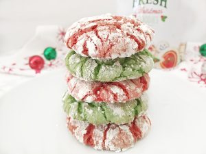 Stack of four baked red and green Christmas Crinkle Cookies heavily dusted with powdered sugar, sitting on a white plate with a festive napkin and ornaments in the background.