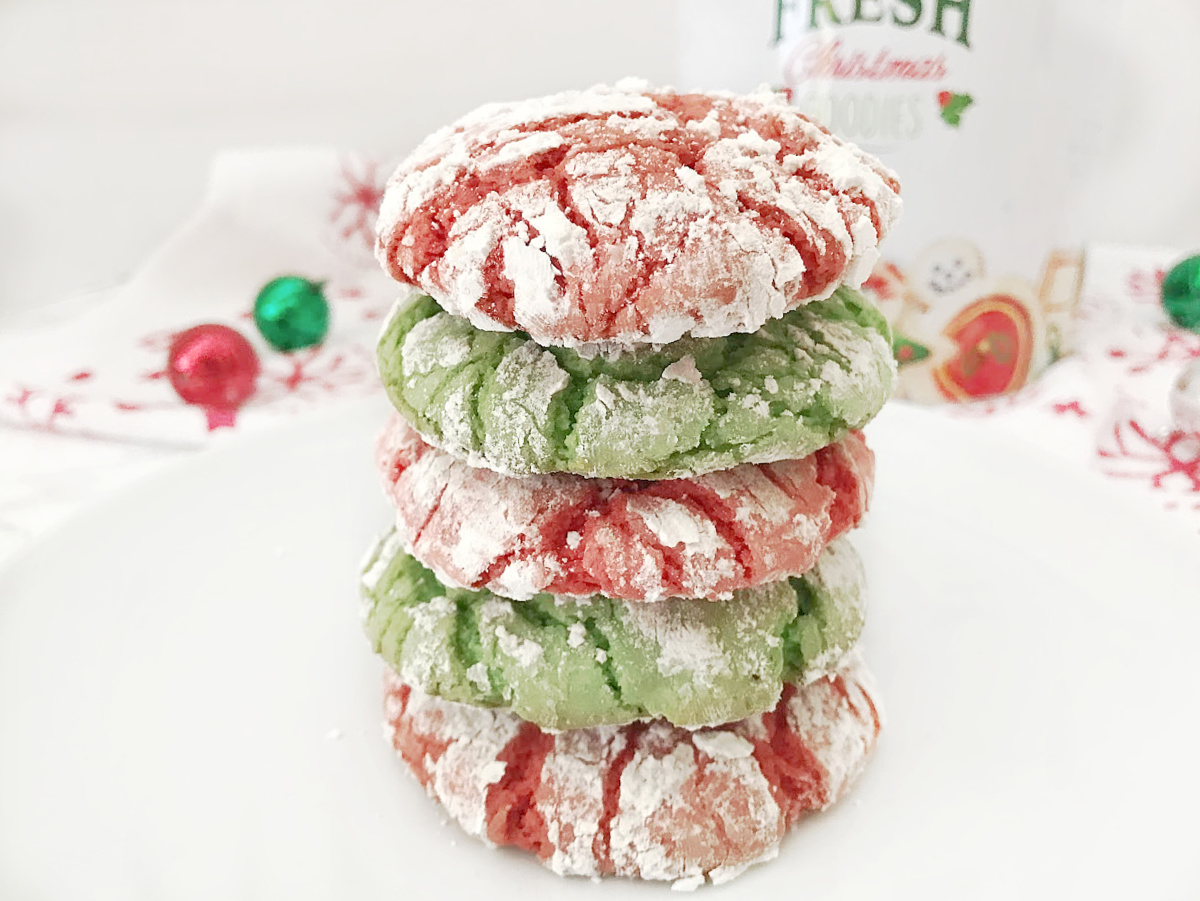 Stack of four baked red and green Christmas Crinkle Cookies heavily dusted with powdered sugar, sitting on a white plate with a festive napkin and ornaments in the background.