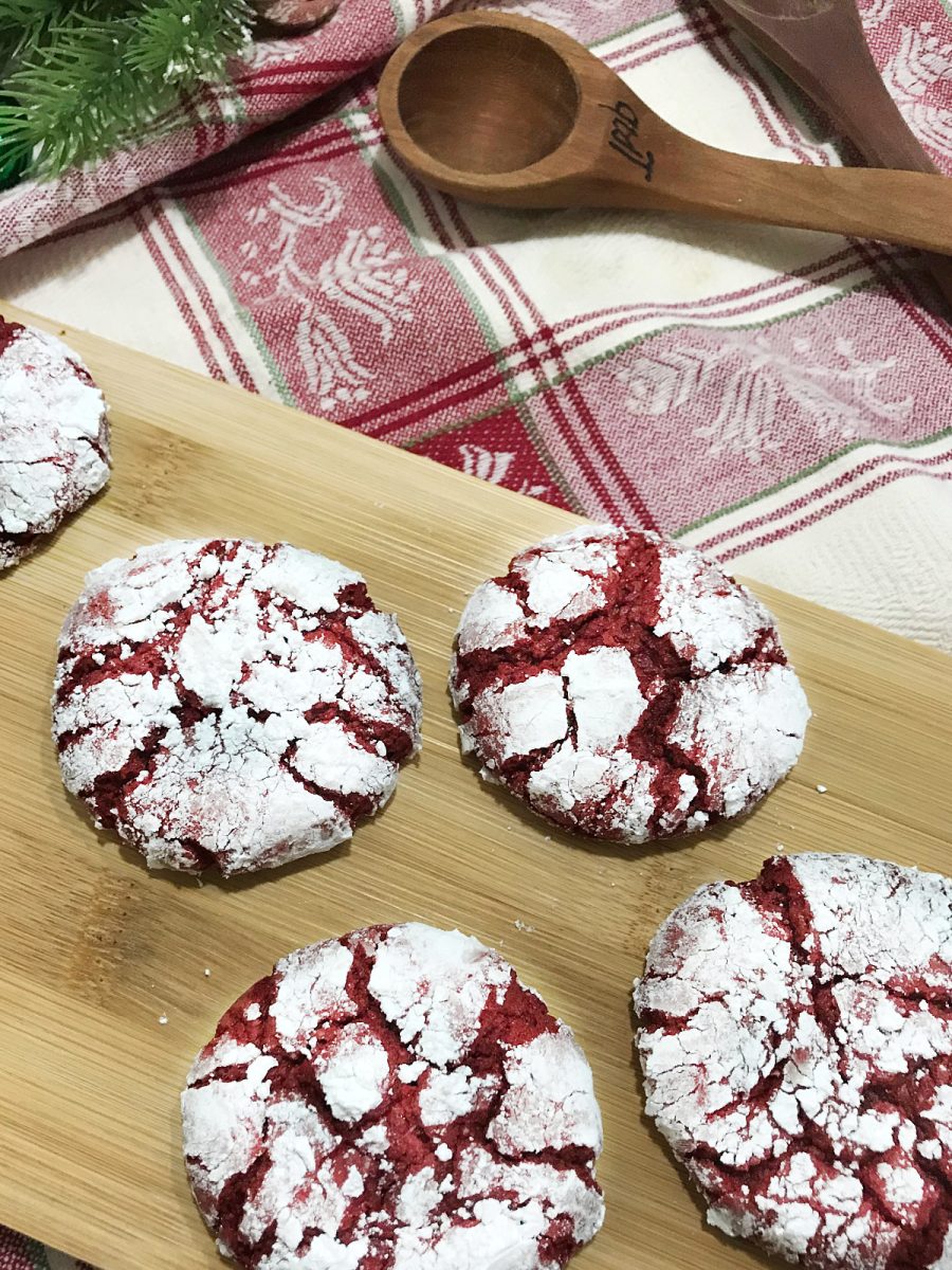 Red Velvet Crinkle Cookies on a wooden board with a festive red and white plaid towel and a wooden spoon.