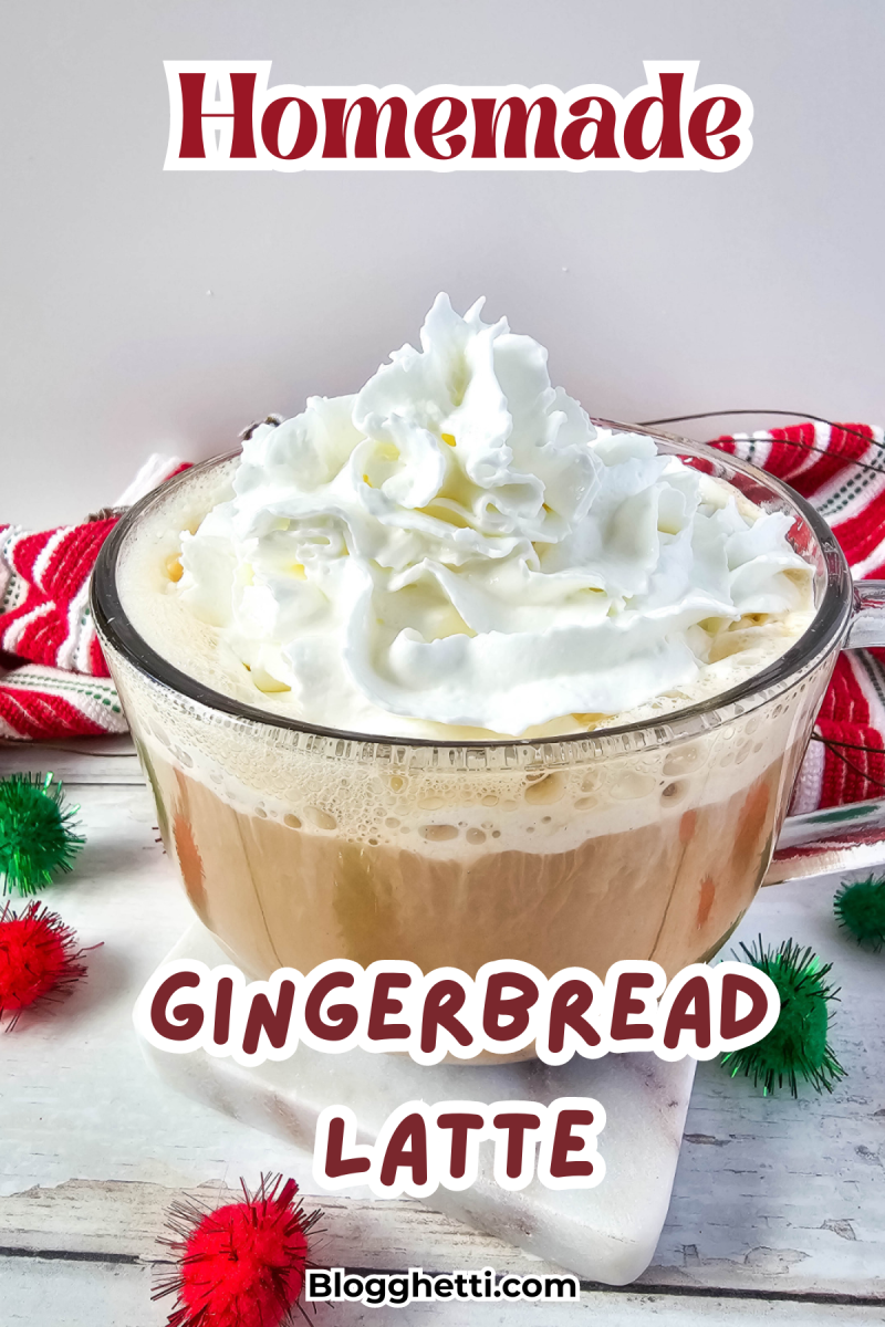 A homemade gingerbread latte topped with whipped cream in a clear glass mug. The latte is surrounded by festive holiday decorations, including colorful pom-poms and a red and white knit cloth, creating a cozy, holiday-themed setting.