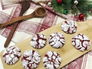 A top-down view of Red Velvet Crinkle Cookies arranged on a wooden board with Christmas ornaments and evergreen sprigs in the background.