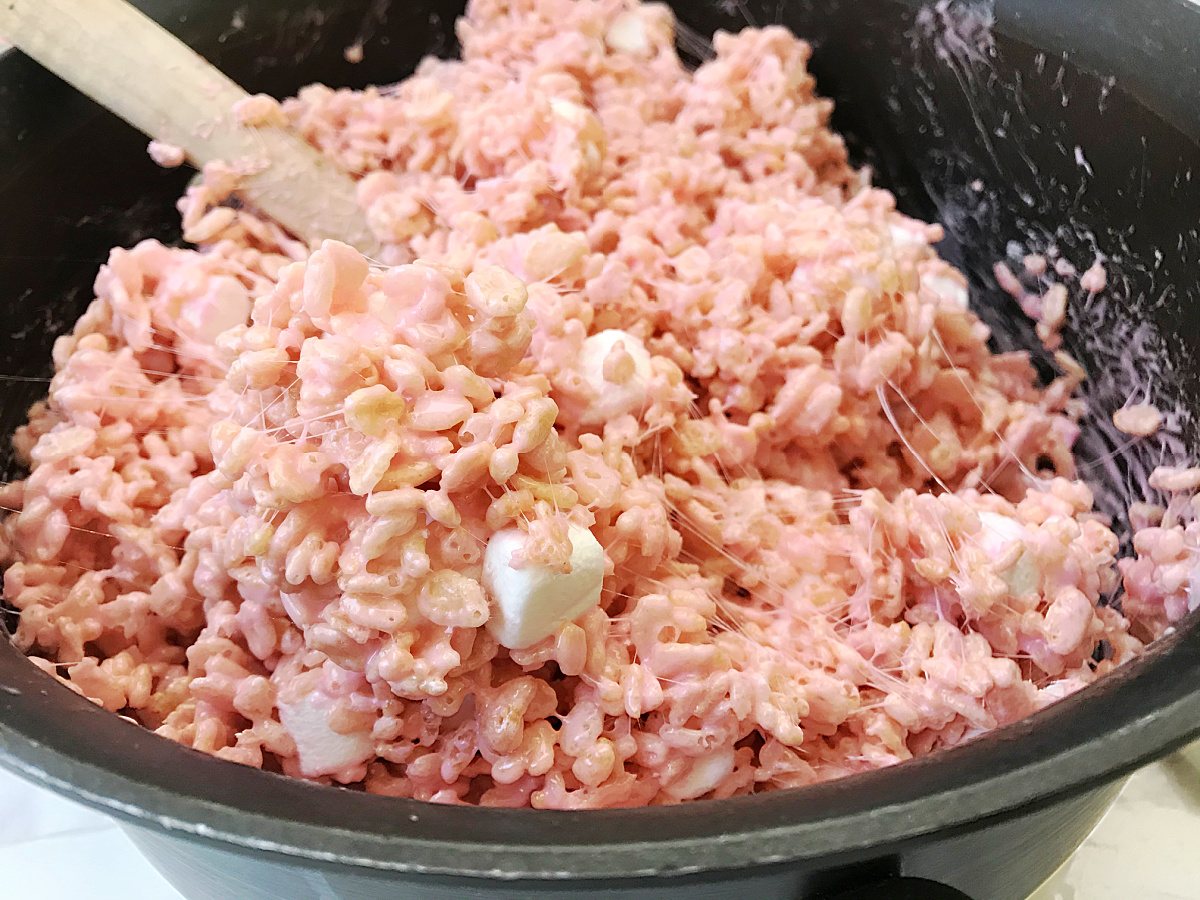 A wooden spoon stirs crispy rice cereal and marshmallows coated in pink mixture, showing sticky strands, inside a dark pot.