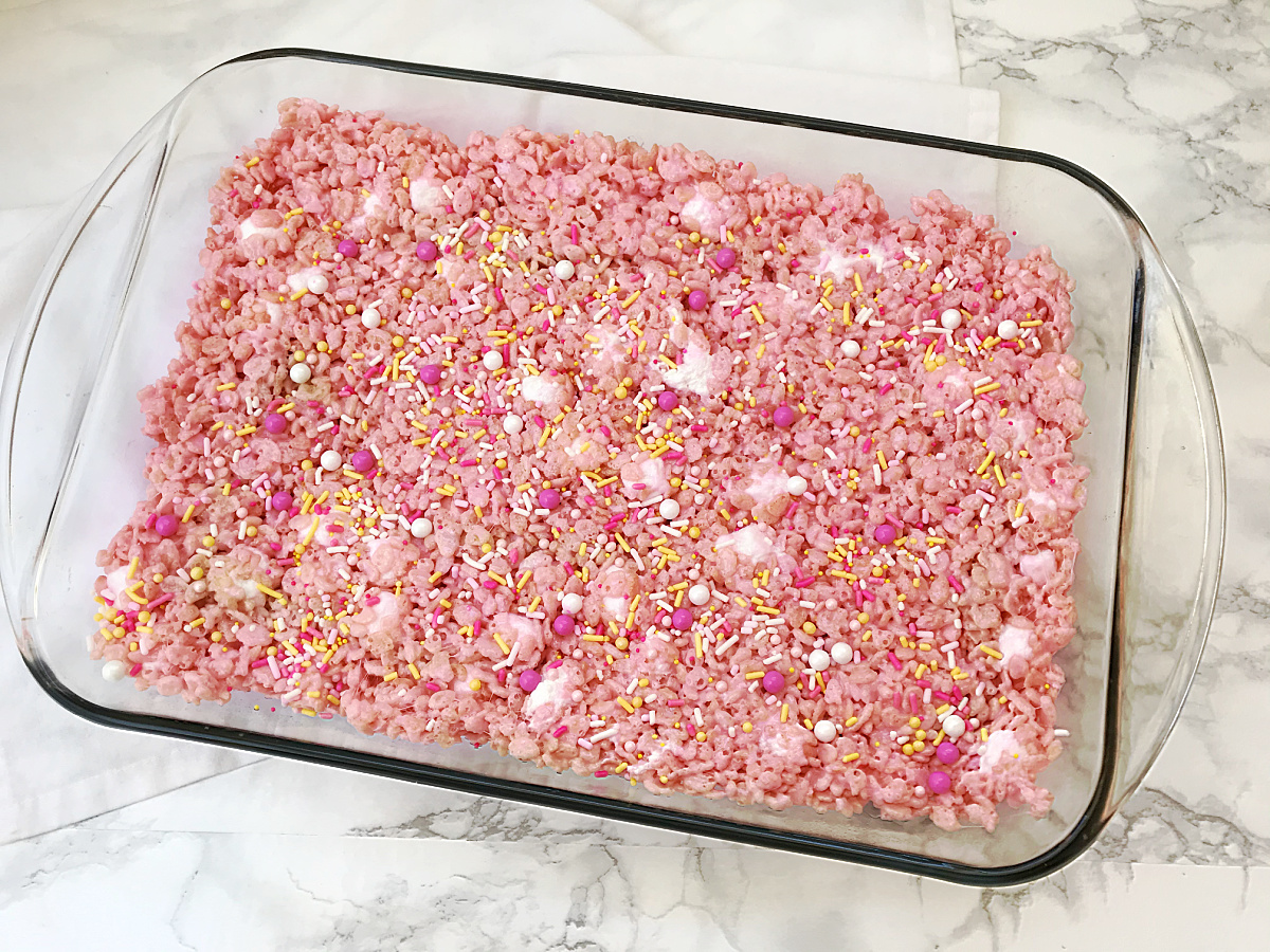 A rectangular glass baking dish filled with pink cereal treats, topped with white, pink, and yellow sprinkles, rests on a white and gray marble surface.