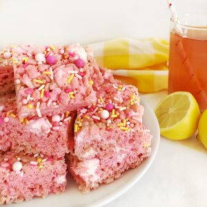 A plate of square pink lemonade cereal treats, topped with white, pink, and yellow sprinkles, next to a glass of pink lemonade and a sliced lemon.