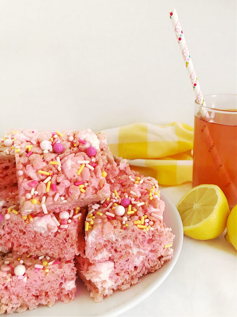 A white plate holds several pink rice krispie treats with colorful sprinkles, positioned next to a glass of pink lemonade with a patterned straw and a sliced lemon.