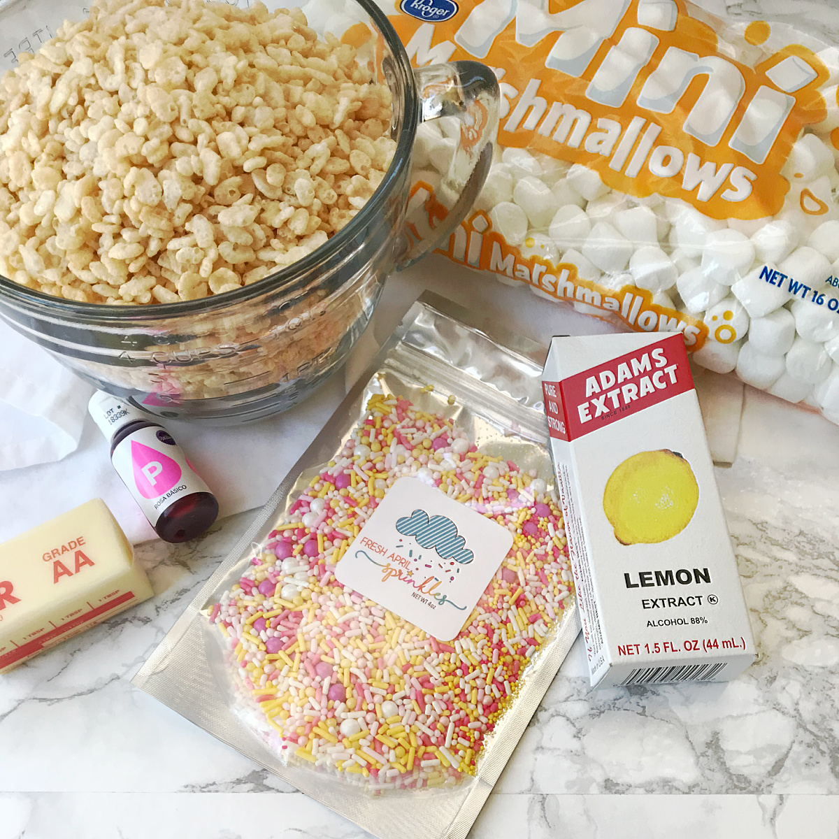 A clear mixing bowl of crispy rice cereal, a bag of mini marshmallows, a stick of butter, a bottle of pink food coloring, a bag of assorted sprinkles, and a box of lemon extract are arranged on a counter.