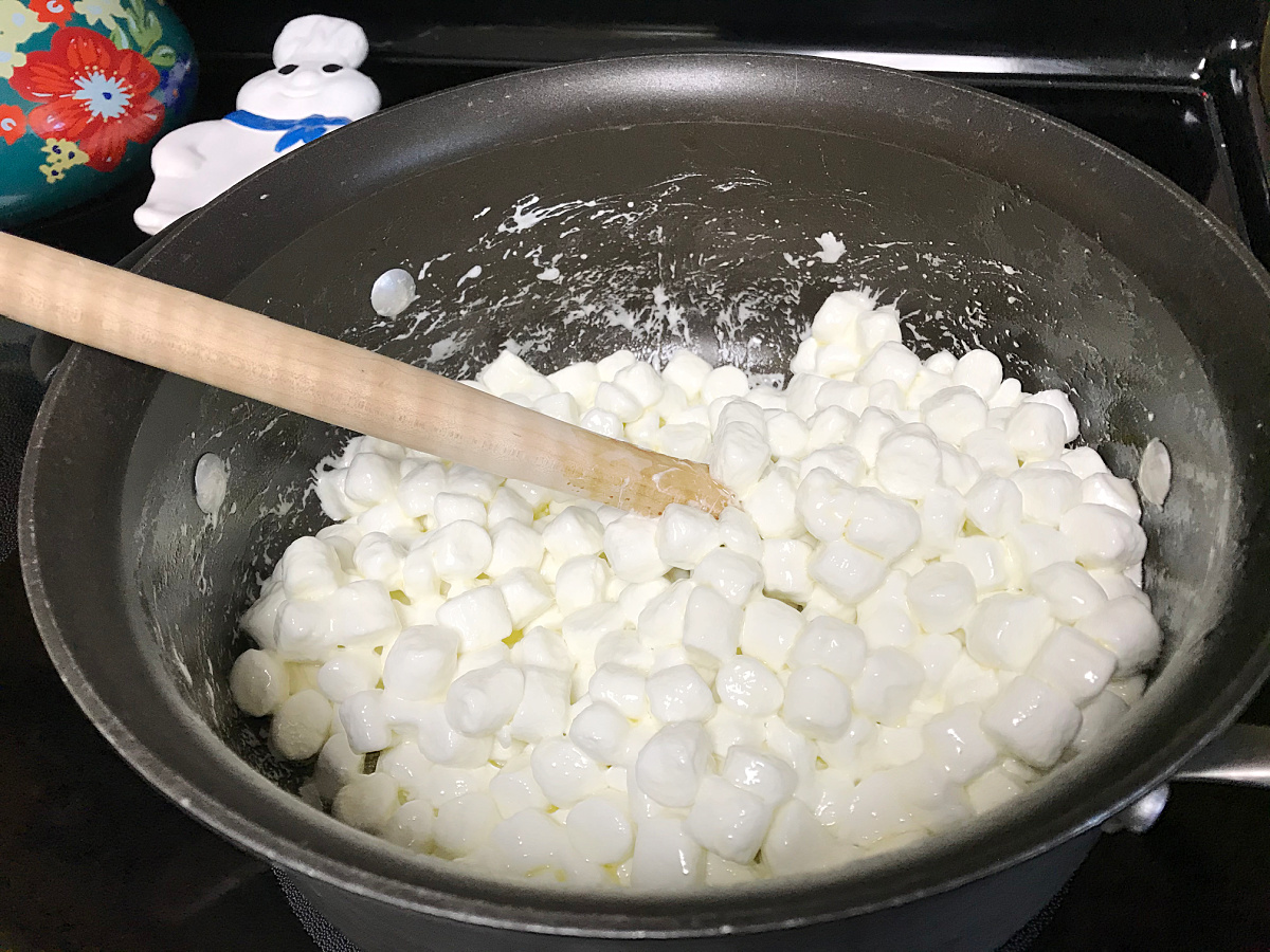 White mini marshmallows are melting in a large black pot on a stovetop, being stirred with a wooden spoon.
