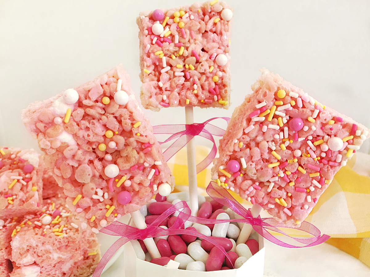 Several pink lemonade treats on white sticks, decorated with pink ribbons and sprinkles, are standing upright in a white container filled with pink and white candies.