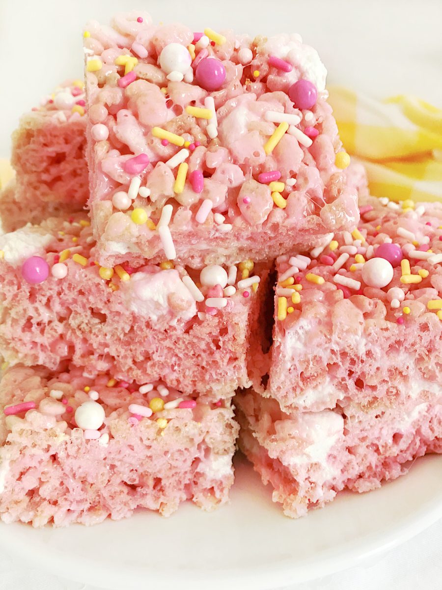 A stack of pink rice krispie treats on a white plate, cut into squares and generously coated with various colorful sprinkles.