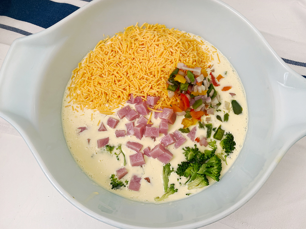 A light blue mixing bowl with a whisked egg mixture, with piles of shredded cheese, diced ham, and broccoli florets waiting to be mixed in.