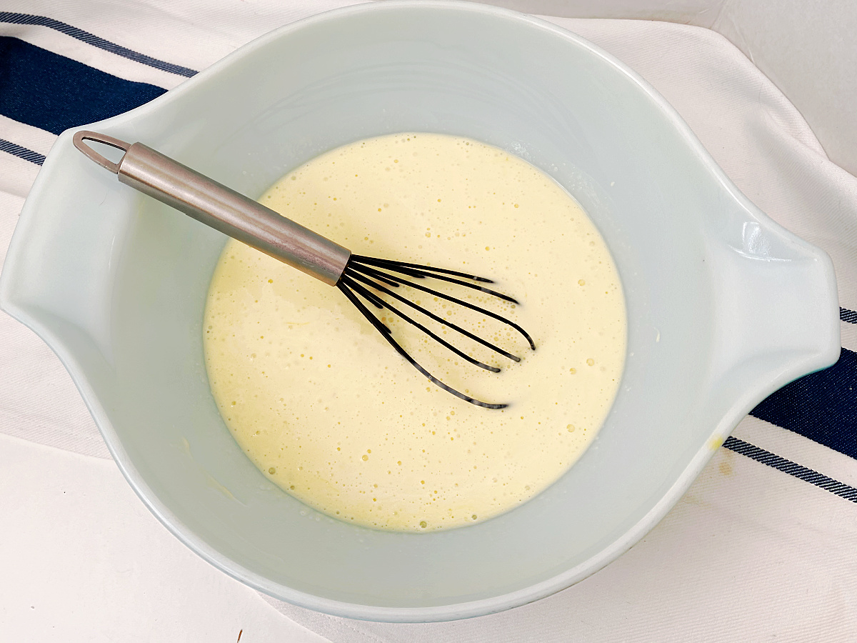 A light blue mixing bowl containing a whisk and a smooth, whisked egg and cream mixture.
