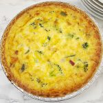 A close-up, overhead shot of a whole baked ham and broccoli quiche in an aluminum pan, showing the golden-brown top and visible ingredients.