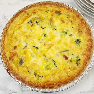 A close-up, overhead shot of a whole baked ham and broccoli quiche in an aluminum pan, showing the golden-brown top and visible ingredients.