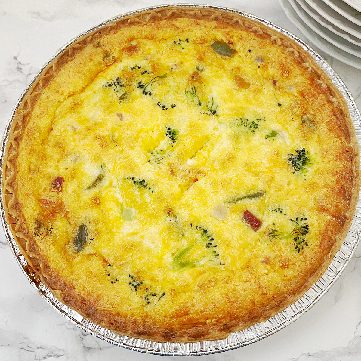A close-up, overhead shot of a whole baked ham and broccoli quiche in an aluminum pan, showing the golden-brown top and visible ingredients.