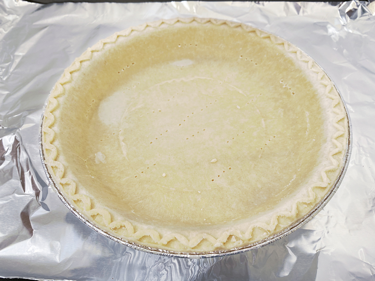 A frozen, unbaked 9-inch deep-dish pie crust in an aluminum pan, with fork marks on the bottom and sides, sitting on a foil-lined baking sheet.