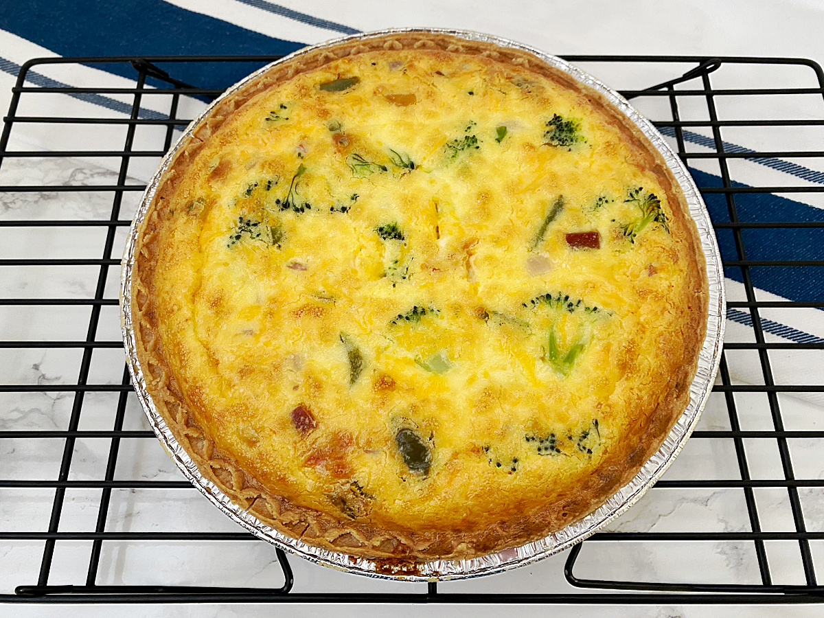 A fully baked ham and broccoli quiche with a golden-brown crust and top, cooling on a black wire rack.