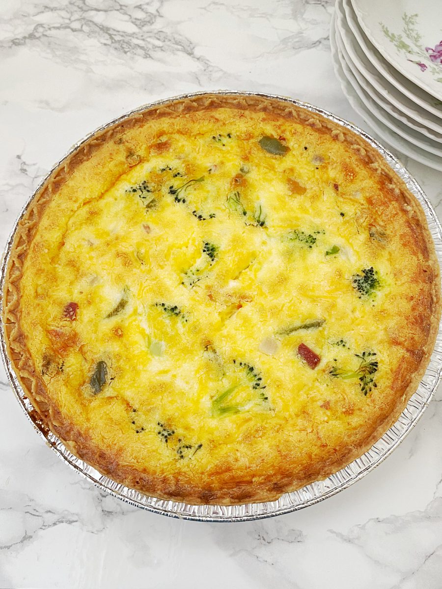 An overhead shot of a golden-brown baked ham and broccoli quiche in an aluminum pan, with a stack of plates in the background.