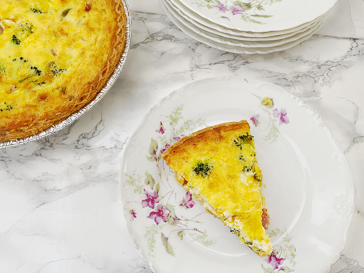 An overhead shot of a slice of baked ham and broccoli quiche on a decorative floral plate, with the full quiche and a stack of plates in the background.
