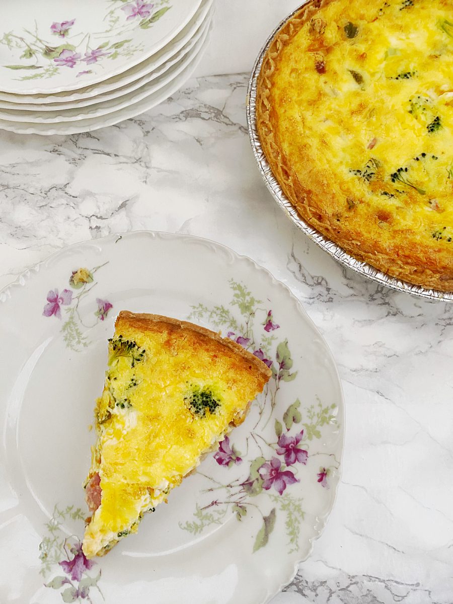 A slice of ham and broccoli quiche on a decorative floral plate, with the full quiche and a stack of plates in the background.