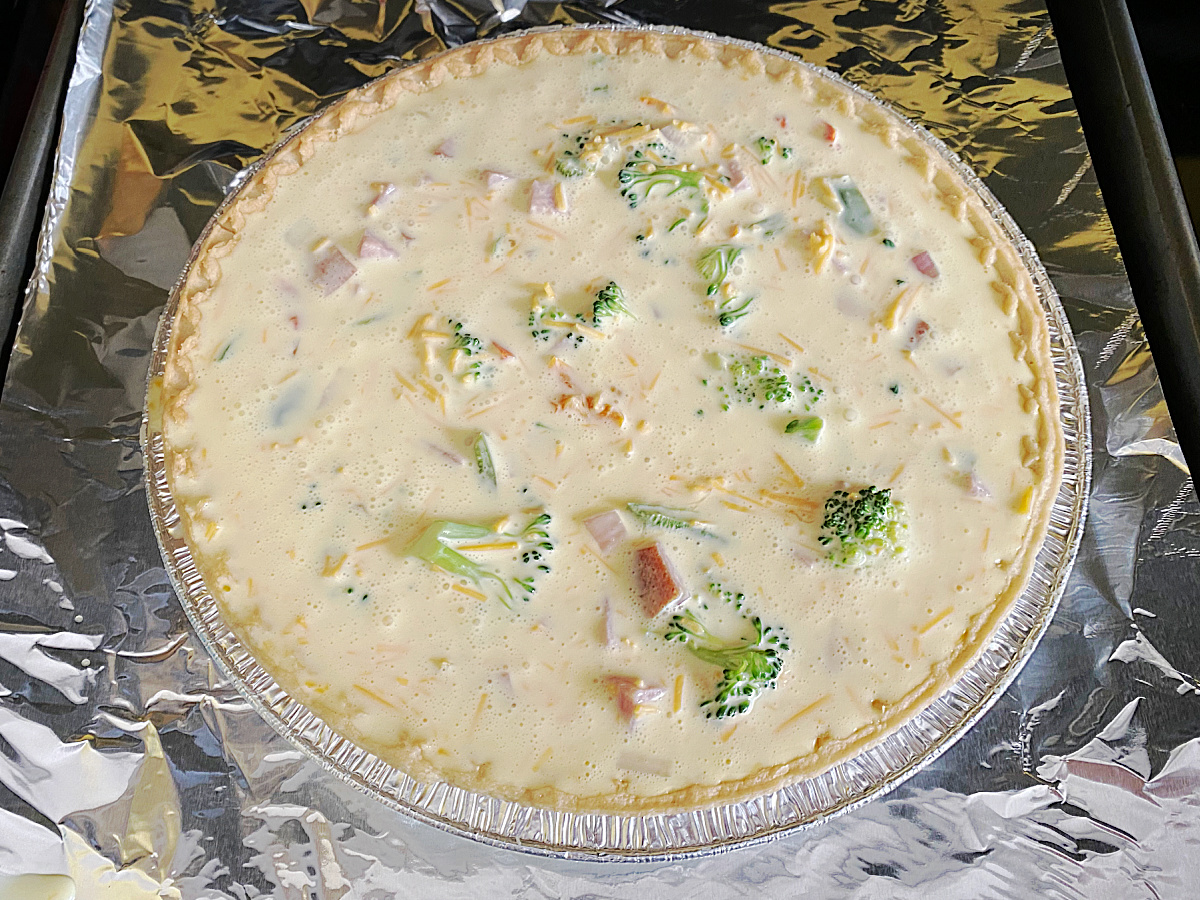 A deep-dish pie crust on a foil-lined baking sheet filled with a creamy egg custard, ham, and broccoli, ready to be baked.