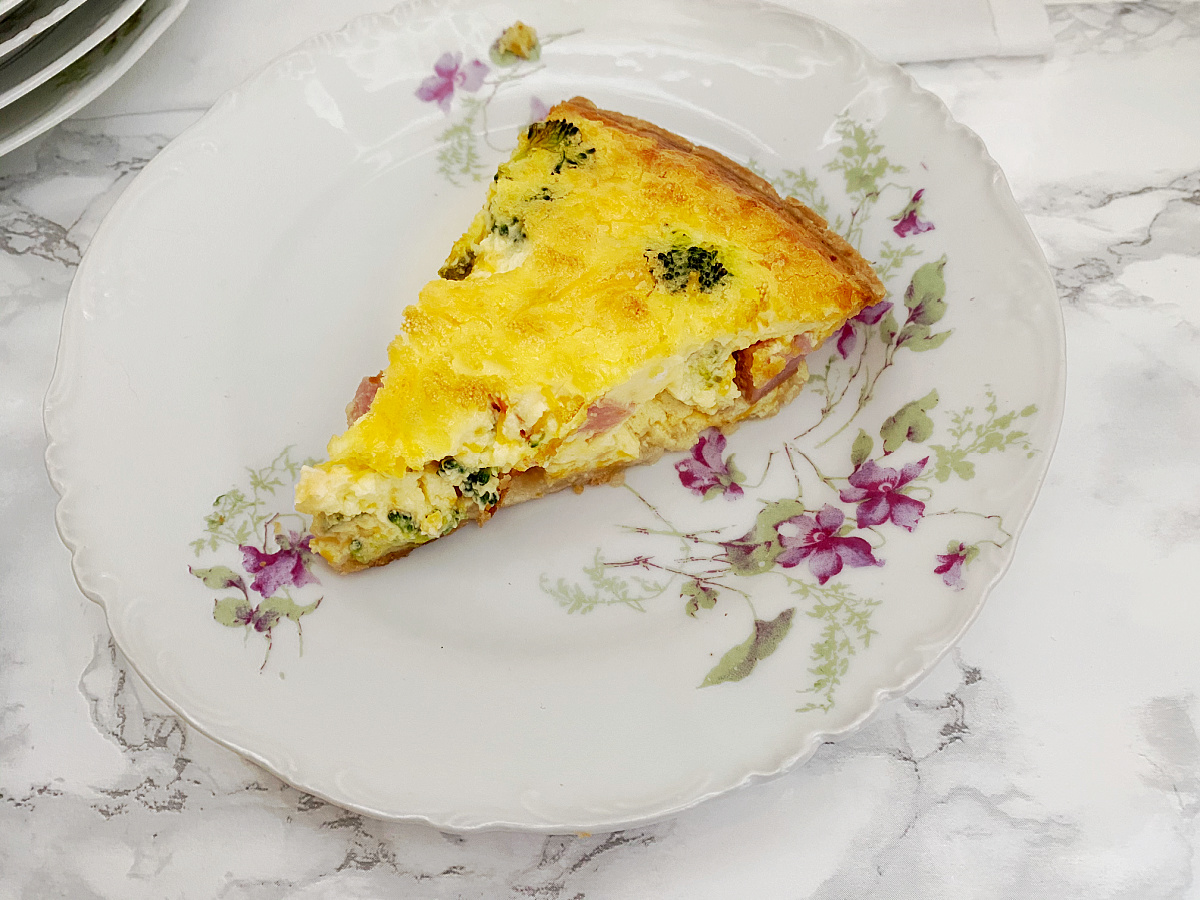 A close-up shot of a slice of baked ham and broccoli quiche on a decorative floral plate.
