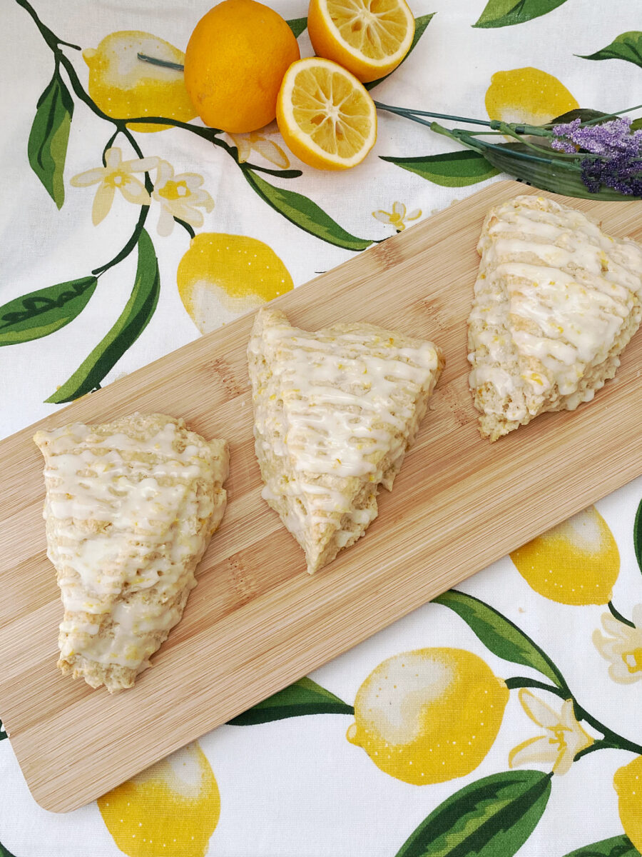 Three golden-brown, triangular scones drizzled with a white lemon glaze and lemon zest, arranged diagonally on a light wooden serving board. The board rests on a patterned tablecloth featuring bright yellow lemons, white blossoms, and green leaves, with fresh whole and halved lemons and a sprig of purple lavender visible in the background.