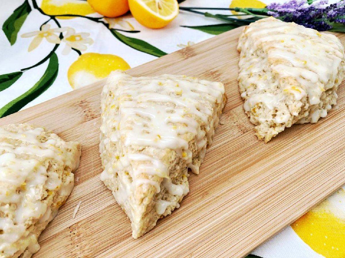 A close-up shot focusing on three glazed lemon lavender scones lined up on a wooden cutting board. The thick, white icing is drizzled generously across the tops, showcasing the textured, crumbly edges of the baked pastry, set against a lemon-patterned backdrop.