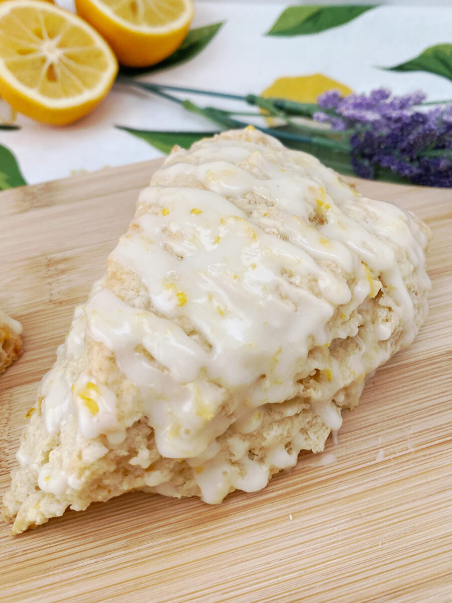 A macro, detailed close-up of a single triangular scone covered in a sweet white glaze and flecks of yellow lemon zest. The scone sits on a wooden surface, with softly blurred fresh lemon halves and a sprig of dried purple lavender flowers in the upper background.