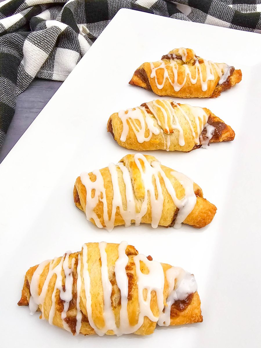 Four freshly baked crescent rolls, filled with a pumpkin mixture and drizzled with white icing, are displayed on a rectangular white plate. A corner of a black and white checkered napkin is visible to the right.