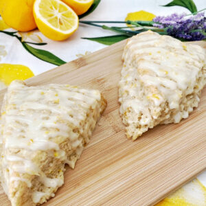 Two freshly baked, triangular lemon lavender scones drizzled with a thick white glaze and lemon zest, sitting on a wooden board. Fresh lemon halves, a whole lemon, and a purple lavender sprig decorate the background on a lemon-themed cloth.