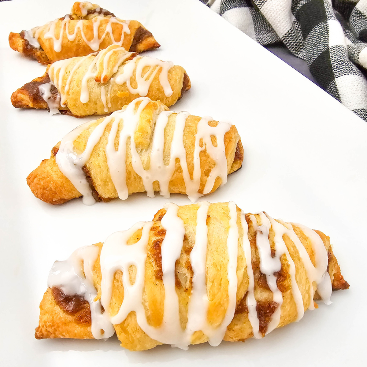 Four golden-brown pumpkin crescent rolls, each topped with a delicate white icing drizzle, are artfully arranged on a sleek white rectangular platter. A black and white checkered cloth is casually draped in the background.