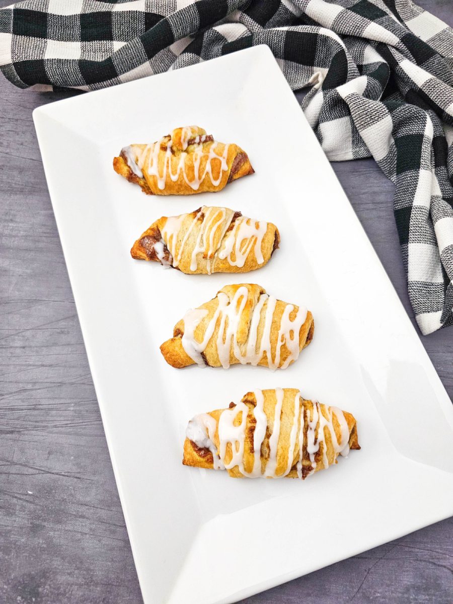 An overhead shot shows four pumpkin pie dessert rolls, glazed with white icing, neatly lined up on a rectangular white serving platter. A black and white plaid dishtowel is in the top right corner against a gray background.